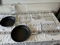 Overview of kitchen lot showing two black non-stick frying pans, three glass casserole dishes including one oval dish with gold rim, and a metal cheese grater with white handle arranged on a tablecloth