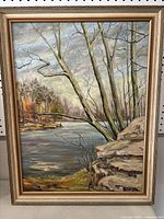 Full view of framed painting showing river landscape, trees, rocks, and sky.