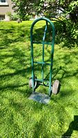 Full view of green metal moving dolly standing upright on grass, showing curved handle, vertical support bars and loading plate.