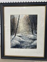 Framed watercolor of a winter forest scene with a stream and snow, in black and gold frame.