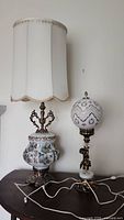Pair of two midcentury table lamps on a wooden surface against white wall, one with fabric shade and ceramic floral base, other with spherical ceramic shade and bronze cherub base.
