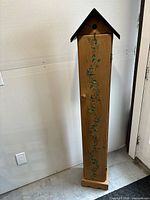 Full view of the tall, narrow wooden cabinet shaped like a birdhouse with peaked roof, front door closed.