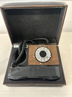Full view of the open portable rotary dial phone showing wood grain dial panel, black handset and coiled cord inside a fitted box.