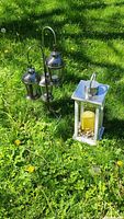 Photo showing both the white rectangular lantern with yellow candle and decorative stones next to the triple silver hanging lantern stand with three lanterns.