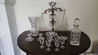 Front view of the balance scale surrounded by crystal glassware including decanter, glasses, vase, and candle holder.