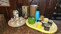 Wide angle showing full lot of vintage bar ware on a marble countertop, including glass decanter, six small hexagonal glasses, green ceramic pot, blue soda siphon, yellow tray with six striped glasses, and four smaller gold etched glasses.