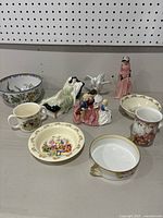 View of all items showing two Royal Doulton figurines, a standing woman figurine, three bowls, and two character mugs.