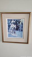 Framed oil painting showing snowy pine trees with light filtering through, artist signature visible.