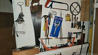 Overview of tools hung on wall including large wooden hand saw, claw hammer, hacksaw, hand level, screwdrivers, pipe wrench, and hanging cage light.
