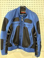 Blue and black Triumph women's motorcycle jacket hanging on white pegboard, showing front view with mesh panels and zippered pockets.