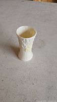 Small off-white Belleek porcelain vase standing upright on a gray surface, showing its textured floral design.
