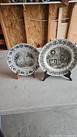 Photo showing two ceramic holiday decorative plates side by side on stands, one with a black and white winter scene and one with a colored fireplace holiday design.
