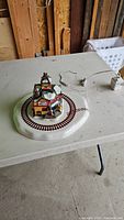 Ceramic Christmas village on a table with train track forming a circle and a ceramic house in the center, with an electrical plug visible.