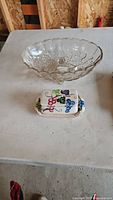Clear glass fruit bowl and ceramic butter dish placed on table; clear glass bowl shows intricate floral patterns and scalloped edges, butter dish lid has grape and leaf decoration.