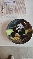 Top side of ceramic collector plate displaying a detailed image of a panda bear sitting and chewing bamboo outdoors.