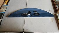 Front view of semicircular wood wall art showing painted design of two loon birds swimming on blue water.