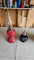 Photo of two oil lanterns, one red metal with glass chimney, and one black ceramic with glass chimney, both decorated with floral patterns.