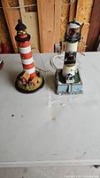 Two decorative lighthouse models placed on a white surface, showing size comparison and power cords.