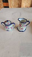 Two-piece porcelain creamer and sugar bowl set with floral pattern and blue handles on a white surface.