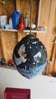 Glass Christmas ornament with hand-painted winter scene featuring an angel in white robe holding lantern and book in a snowy landscape. Hanging cord attached.
