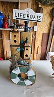 Wooden nautical-themed decorative item with birdhouse shapes, rope, painted lifebuoy ring, blue bird figurines, decorative leaves, and a sign that says 'to LIFEBOATS'.
