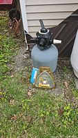 Side view of the gray cylindrical sand filter tank with black multiport valve on top and assorted hose connections, placed outdoors on grass with nearby shed wall.