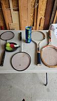 Full view of all rackets, can of tennis balls, and shuttlecock pouch on white table.