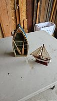 Two wooden nautical model boats on a gray table. One is an open boat frame painted green inside, the other a sailboat with sails mounted on a stand.