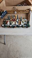 Wooden Christmas decoration featuring painted snowmen figures, pine trees, pinecones, and 'Let It Snow' sign within a wood stick frame with hanging twine.