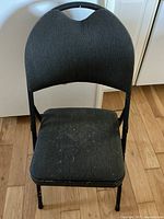 Front view of folded black chair showing padded striped fabric seat and back with metal frame and visible wear.