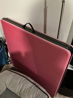 Folding table folded, showing its faded red plastic top with a black carrying handle on top.