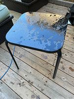 Black metal outdoor side table shown on wooden deck, reflective top surface with visible wear and dirt from outdoor exposure