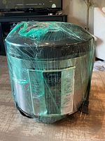 Front view of the Midea multifunction cooker wrapped in plastic wrap, showing control panel and attached power cord.