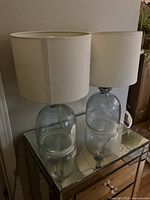 Two modern table lamps with glass bases and white cylindrical fabric shades on a mirrored surface.