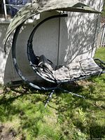 Outdoor swing lounge chair with gray cushion and canopy shown assembled outdoors on grass near a wall.