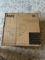 Image of RMS branded cardboard box featuring a line drawing of the elevated toilet seat with padded arms and the text indicating it is a universal elevated toilet seat.