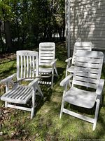 Photo showing four white plastic outdoor chairs with horizontal slats, arranged on grass near a building, under sunlight.