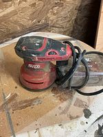 Red and black Skil 7492 orbital sander with power cord and dust collection box on wooden surface.
