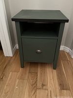 Front view of a small dark green painted side table with one open shelf and a drawer with a metal handle.