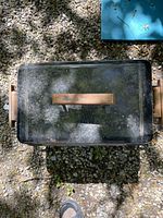 Top view of the rectangular black metal portable gas grill with a wooden handle on the lid placed on gravel outdoors.