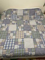 Full view of the quilt spread on a bed, showing multiple plaid patchwork squares in blue, green, beige, and white tones.