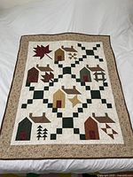 Full view of the handmade lap quilt with visible patchwork design of houses, leaves, and trees on a cream background with beige floral border.