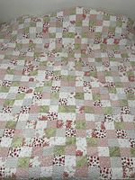 Comforter with patchwork squares in pink, green, and white draped over a bed, showing full view of pattern and texture.