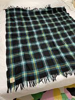 Full view of the black Campbell tartan wool blanket spread on bed, showing the blue, green, yellow, and gray plaid pattern and fringed edges.