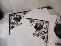 Pair of metal shelf brackets with rooster decoration and swirled scroll patterns placed on white background.