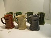 Front angle view of 7 ceramic mugs of various colors including green, brown, cream and blue with Alexander Keith's logo embossed on them.