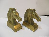 Pair of brass horse head bookends positioned side by side, front view. Shiny finish with detailed horse manes and facial features.