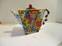 Side view of the tea pot showing colorful geometric and cartoon pattern, red knob, zigzag handle, and dotted spout.
