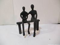 Front view of two black cast iron stylized human figures sitting closely on a bench. The figure on the right has an arm placed behind the other figure's back.