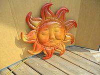 Front view of metal sun wall hanging resting on wood deck against cardboard background. Shows detailed facial features and orange-yellow gradient with curved sun rays.